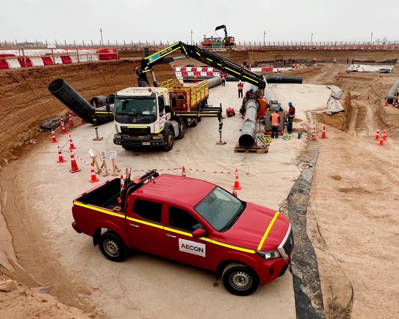 Camioneta roja de AECON y camión pluma junto a tuberías en una excavación de faena